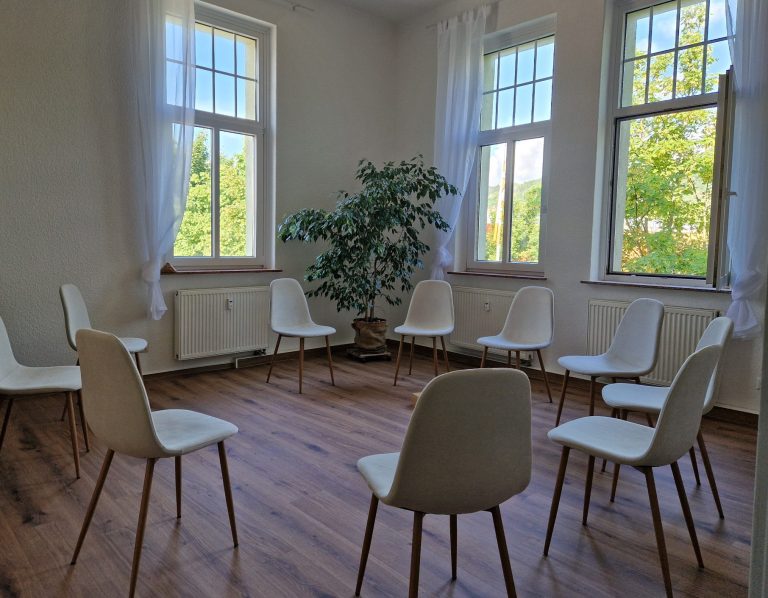 Ein heller Raum mit Stühlen im Kreis und einem Pflanzenarrangement in der Ecke.