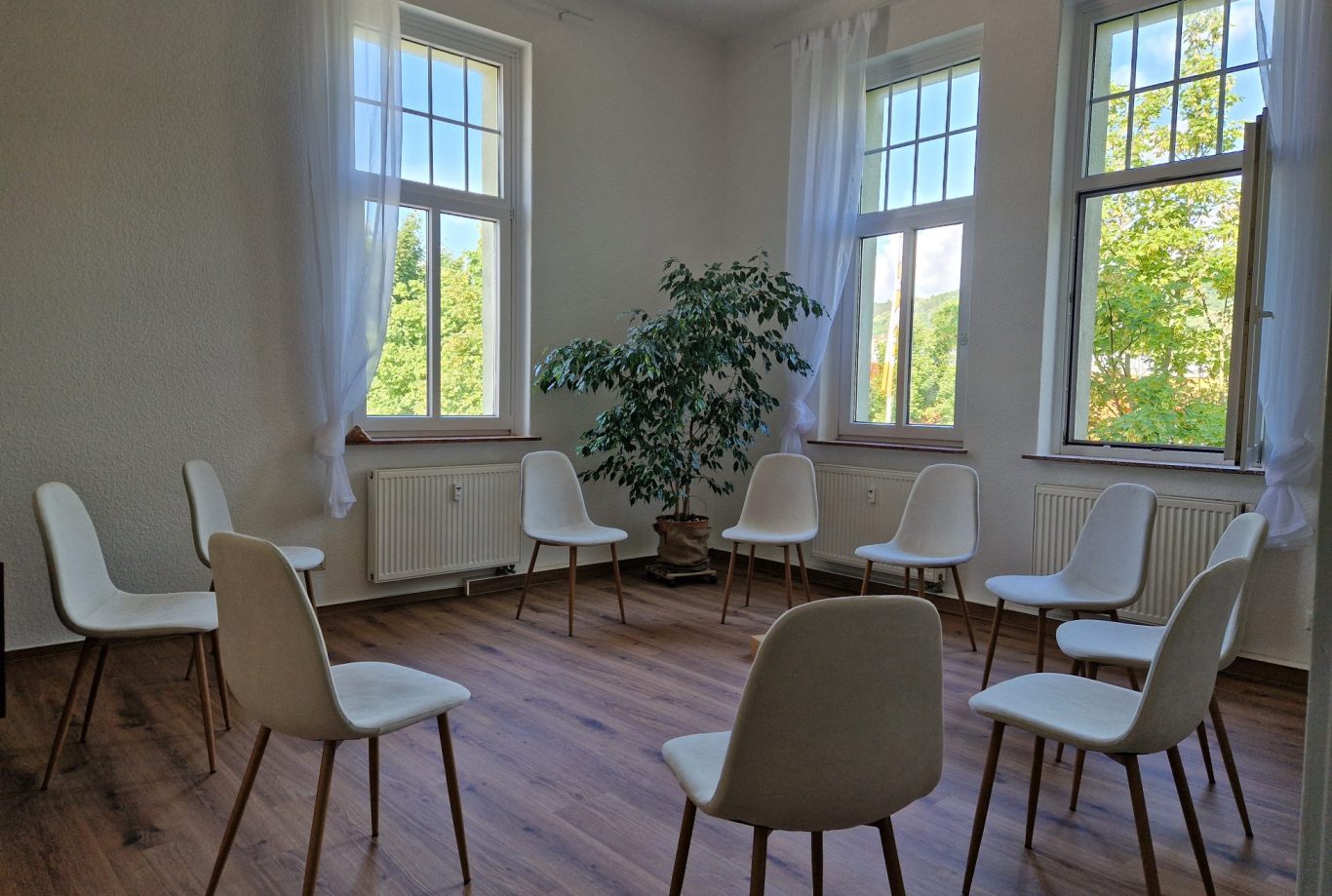 Ein heller Raum mit einem Stuhlkreis und einem großen Fenster. Pflanzen sind sichtbar.